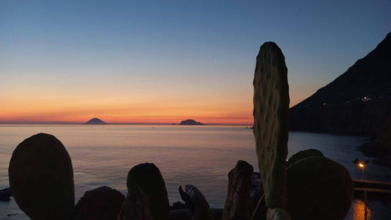 Tramonto sul mare con il profilo delle isole all’orizzonte e gli scogli in primo piano