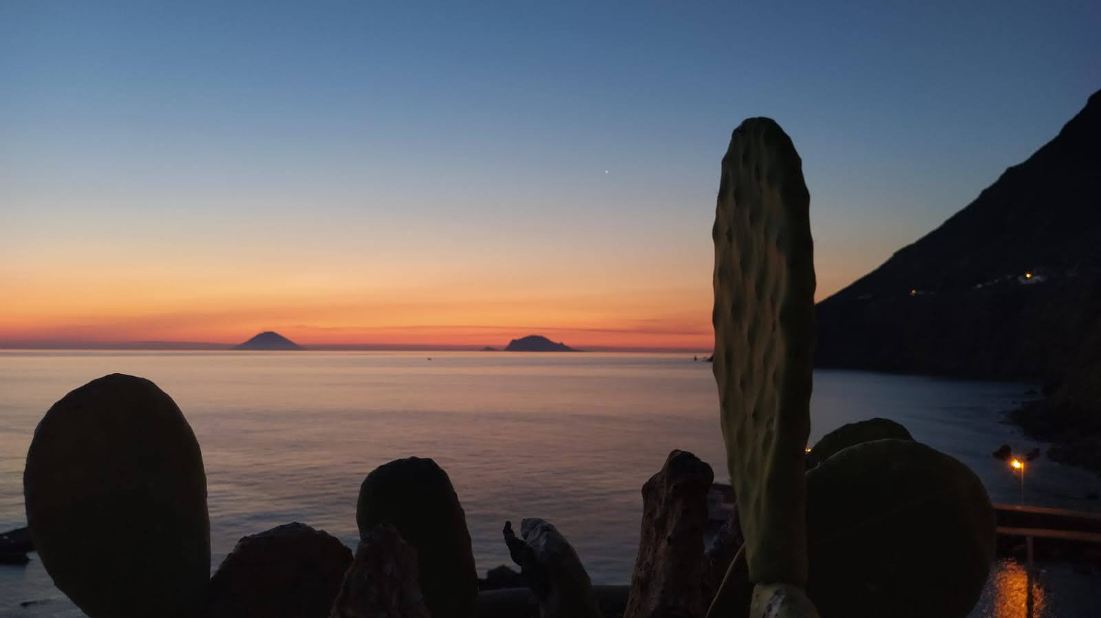 Tramonto sul mare con il profilo delle isole all’orizzonte e gli scogli in primo piano